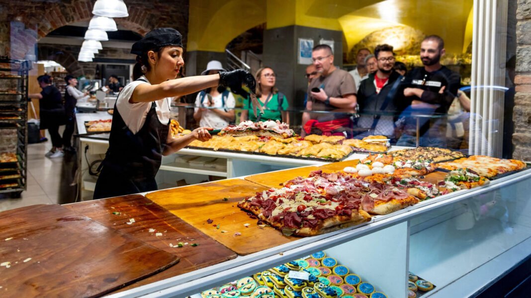 A female restaurant worker slices pizza slices for customers behind a display case in a small paninoteca and pizzeria shop in the Bergamo Italy.