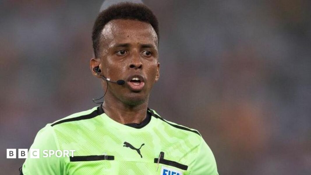 Omar Artan refereeing a match, with earpiece and microphone, and a Fifa badge on his light green shirt