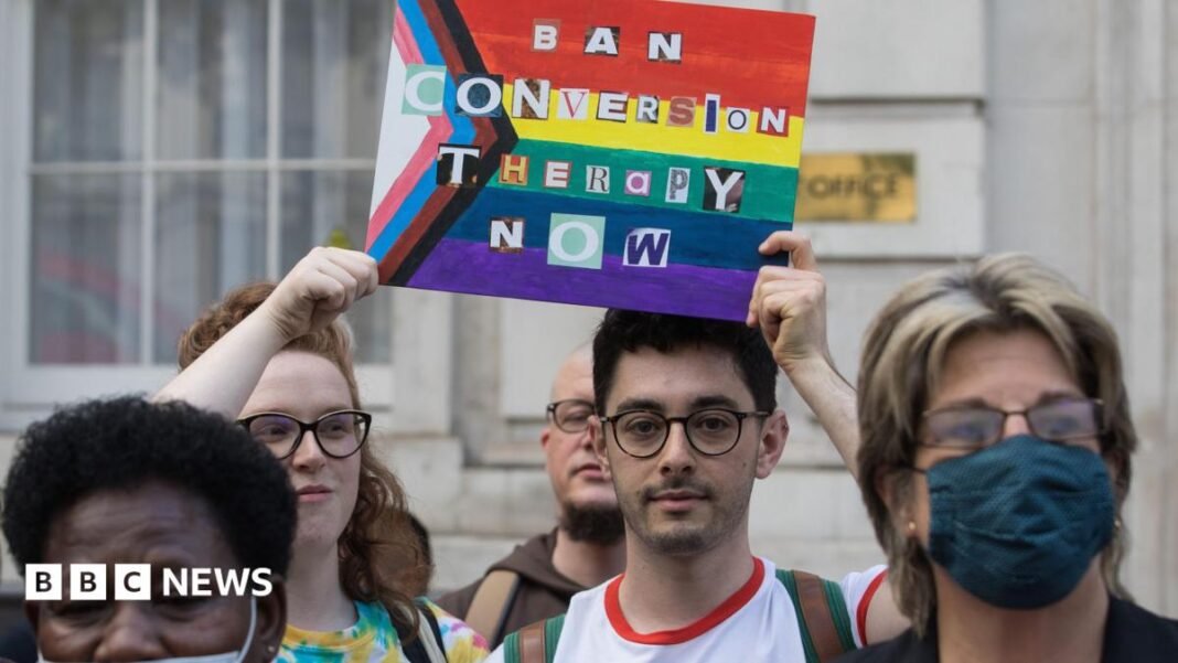 A tightly cropped image of campaigners protesting for a ban on conversion practices.  Two campaigners are in sharp focus, holding a sign featuring the progress pride flag and the words 'Ban Conversion Therapy Now' written in letters cut out from a newspaper or magazine. Both protestors are wearing glasses and have serious looks on their faces. Other protestors in the foreground are slightly out of focus.
