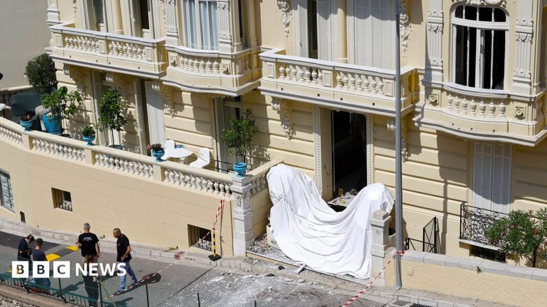 Frontage of a large cream coloured residential building in Monaco with damage to windows and a white sheet accross the front steps and police standing near the front of the building