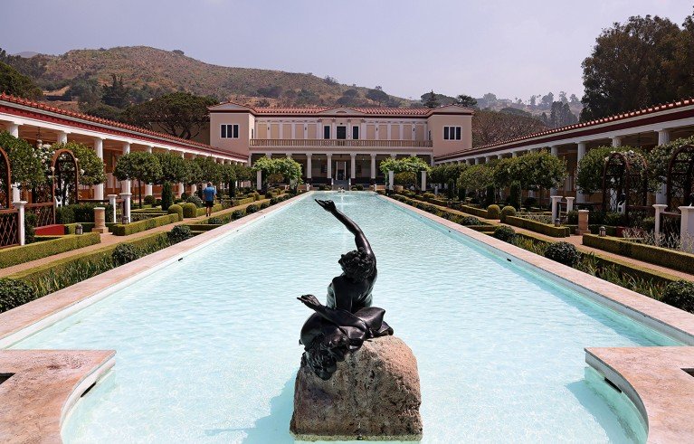 Long rectangular reflecting pool with a bronze statue of a reclining figure on a rock in the centre foreground, flanked by manicured gardens and arched trellises. A large classical-style building with columns and a red-tiled roof is visible at the far end.