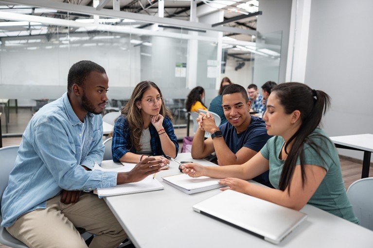 A small group of students sit around a long table, actively discussing ideas in an academic setting while their laptops and notebooks remain unused.