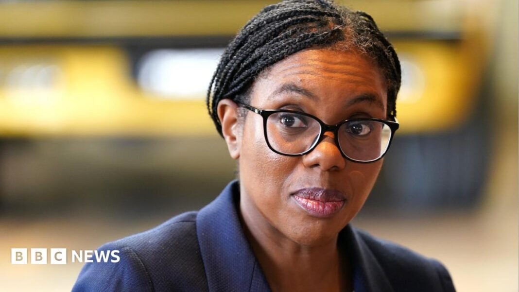 A picture of Kemi Badenoch from the shoulders up. She is wearing a dark blue suit jacket and black-rimmed glasses. Her hair, which is in braids, is tied back. She is looking to the left and the background is blurred.