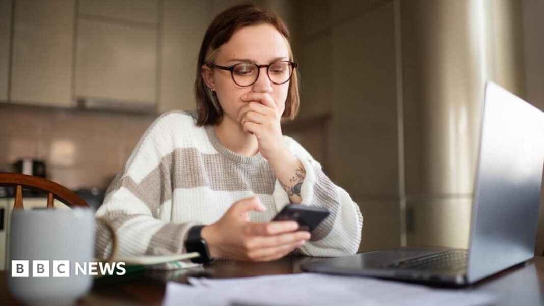 A young woman with round glasses and short brown hair wearing a striped white and beige long sleeved top and a smart watch is sat at a kitchen table holding her smartphone with her laptop open, papers in front of her, and tea by her side. She appears to be struggling to find job opportunities online and her hand is over her mouth.