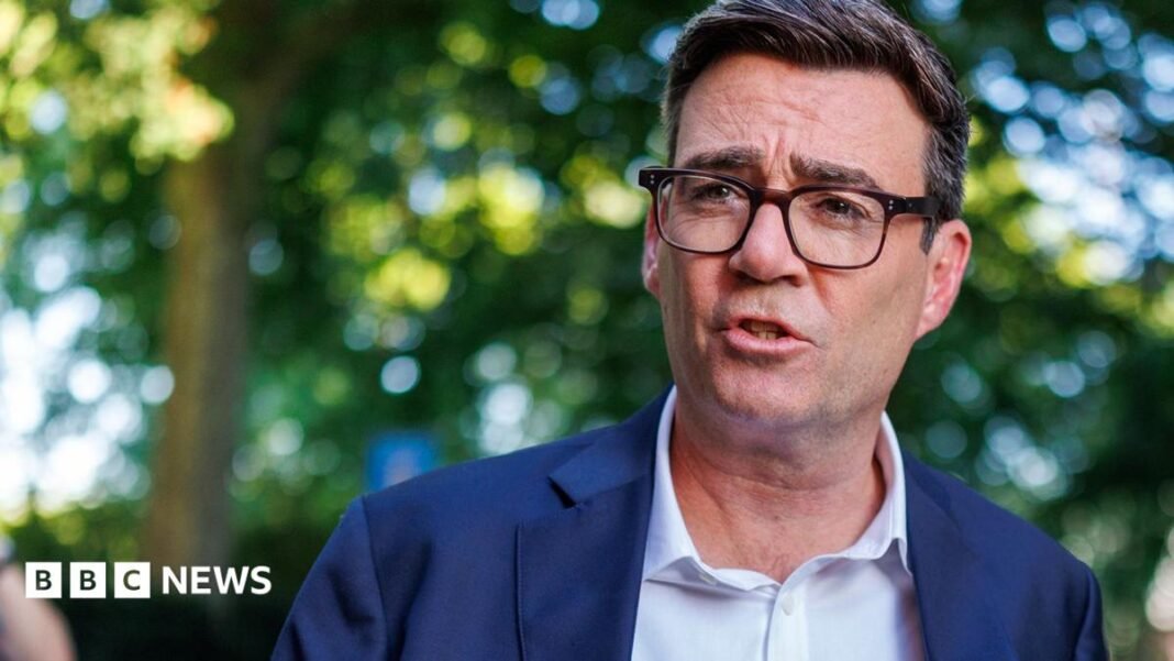 Andy Burnham, wearing a suit, white collared shirt and glasses. Trees in sunlight can be seen in the background