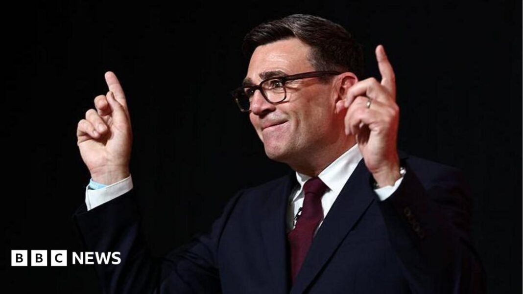 Andy Burnham, in a dark suit and red tie, raises both hands to point up, as he delivers his leadership 