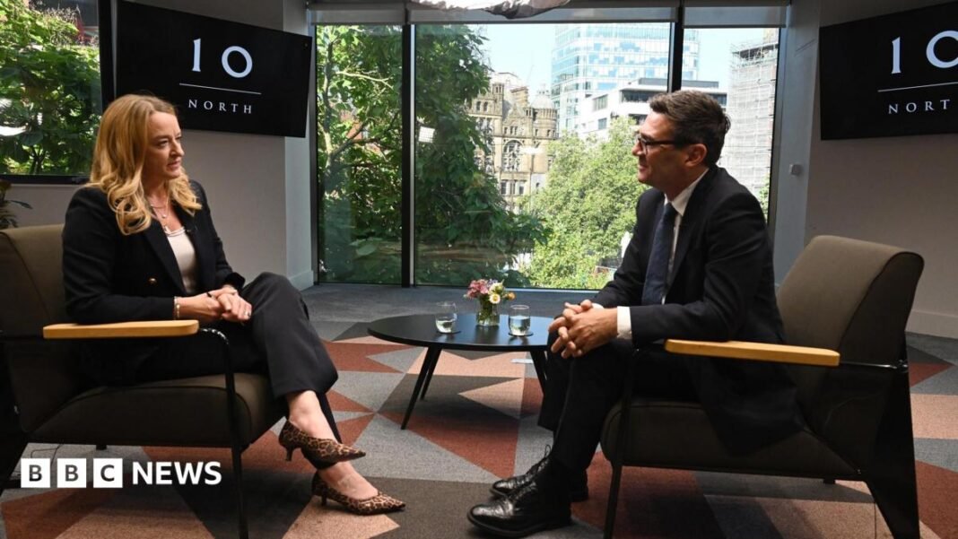 Andy Burnham, wearing a dark suit, gestures with his hands while speaking during an interview. Behind him is a TV with the logo for No 10 North.
