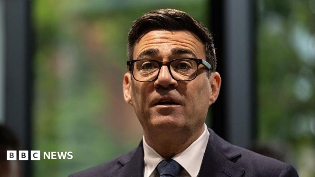 A picture of Andy Burnham from the shoulders up. He is wearing a dark suit with a white shirt and dark tie, has thick-rimmed black glasses on, and has short, dark brown hair. The background behind him is blurred and he is looking forward with his mouth slightly open and his eyebrows raised.