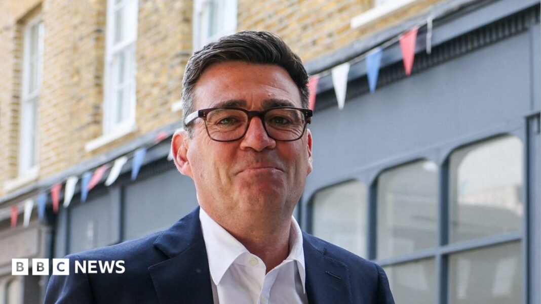 Andy Burnham looks directly forward and smiles with his mouth closed. He wears tortoise shell glasses, a navy suit jacket is navy and a white shirt. He stands in a street, behind him is a blue shop, with red, blue and white bunting hung above it.