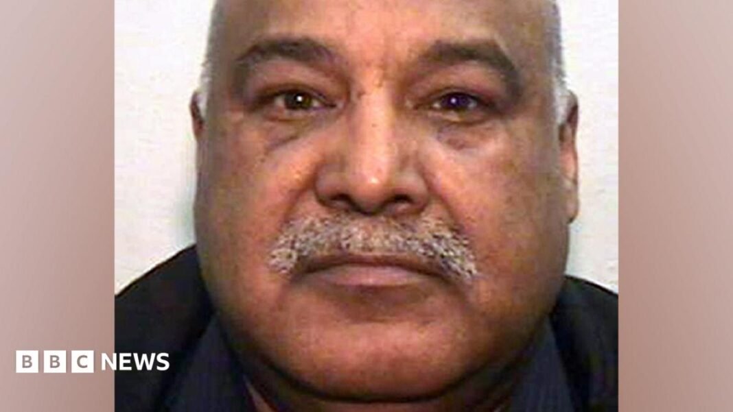 A mugshot of an Asian man in his 60s, with a grey moustache and bald head. He is looking into the camera.