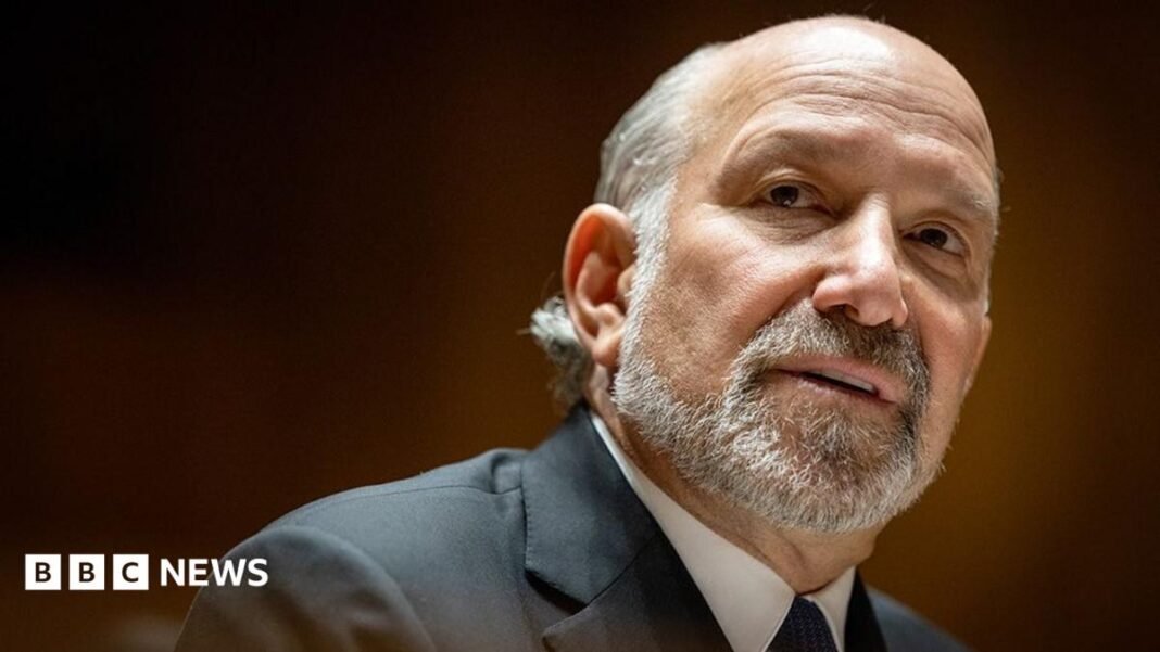 Close-up portrait of Howard Lutnick with a short grey beard and receding hairline, wearing a dark suit jacket, white shirt, and dark tie. The person is turned slightly to the side and looking toward the camera. The background is softly blurred with warm brown and amber tones, creating a shallow depth-of-field effect.