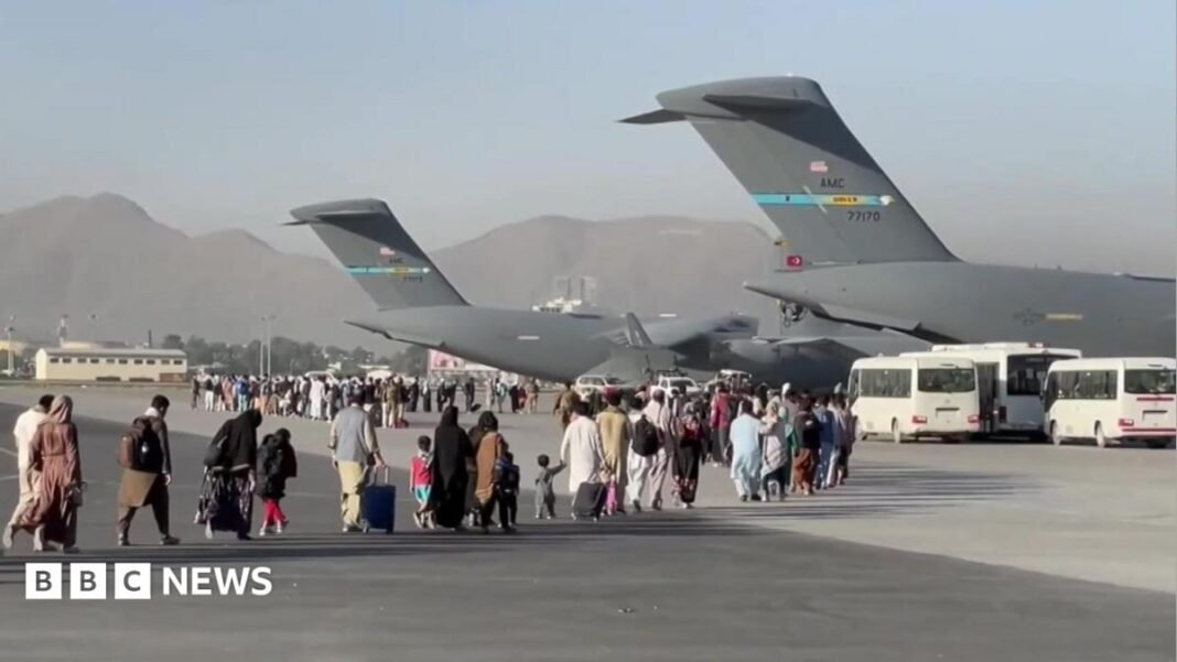 People carrying luggage walk across an airport tarmac towards large US military transport aircraft during an evacuation