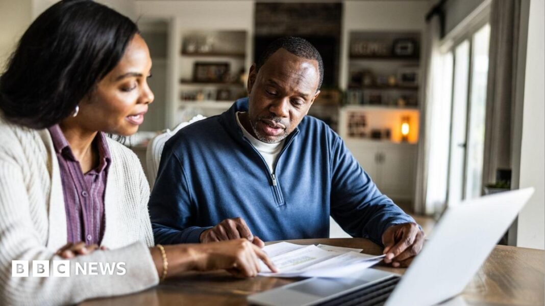 Middle-aged couple look at bills on a table in front of a laptop, with a living room out of focus behind them.