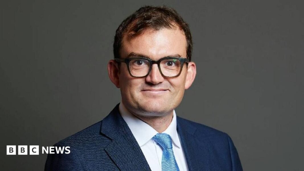 MP found not guilty of sexually assaulting women Patrick Spencer wears a navy suit and stands in front of a plain background. He is smiling directly at the camera and is wearing large black-rimmed glasses.