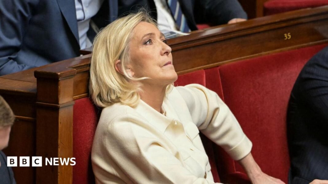 Marine Le Pen sits in France's National Assembly in a red velvet seat. She is biting her lip and looking up. She wears a cream suit. 