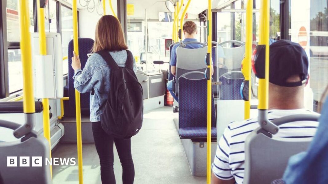 Most bus fares in England to be capped at £2 A young woman wearing a yellow jacket and pink headphones is holding a mobile phone while sitting on a bus.