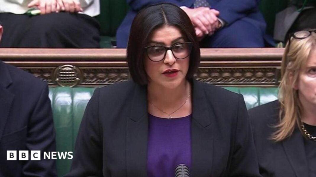 Mahmood wears a black jacket over a royal purple top. She wears her black hair in a chin length bob, with spectacles and red lipstick.