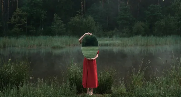 fair-skinned person in a red dress holds up an oval mirror in front of their face and torso while standing outside