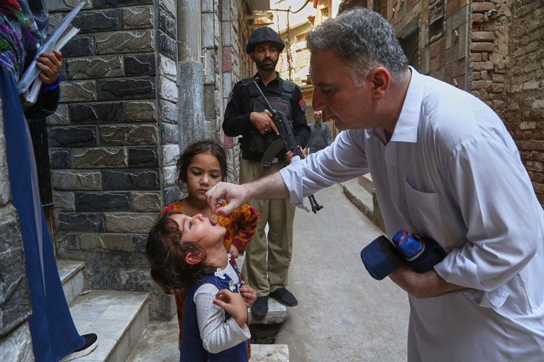 On the steps of a doorway, a health worker administers a polio vaccine to a child as an armed police officer stands guard.