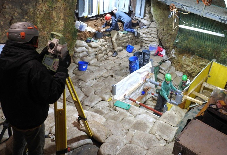 People in hard hats work in a trench reinforced with sandbags.