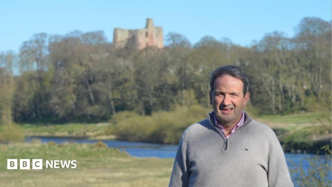 Northumberland County Councillor Patrick Lambert. He has short salt and pepper hair and is wearing a grey jumper. He is standing in an open area surrounded by grass, with a stream and a castle ruin in the background.