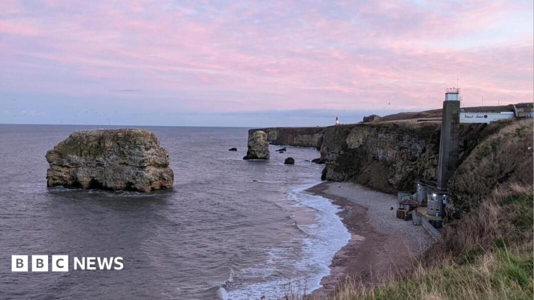 Council cancels climate emergency declaration Purple clouds over Marsden Rock, a large rock surrounded by the sea. It is a short way away from a beach, where there is a restaurant at the bottom with a large concrete lift shaft leading to the top