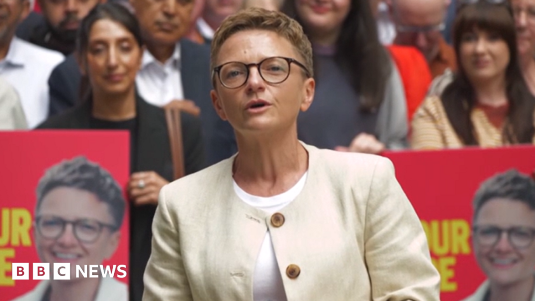 Bev Craig, wearing a white jacket, and in mid-speech. She has short blonde hair and is wearing glasses. There are people sitting behind her, as well as posters with her face on them.