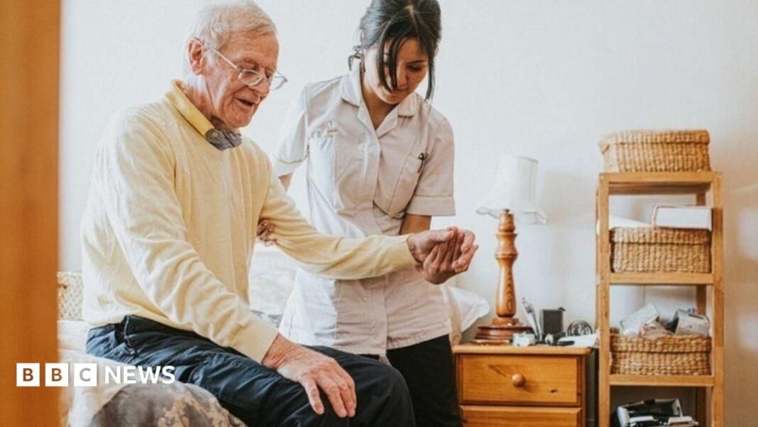 An older man with a younger nurse, who is holding his left hand. Some bedroom furniture is pictured to the left of him.