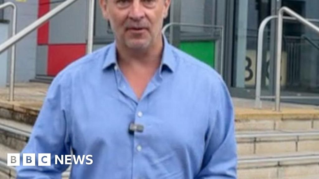 A journalist wearing a light blue shirt and a microphone stands outside a BBC building in the background. 